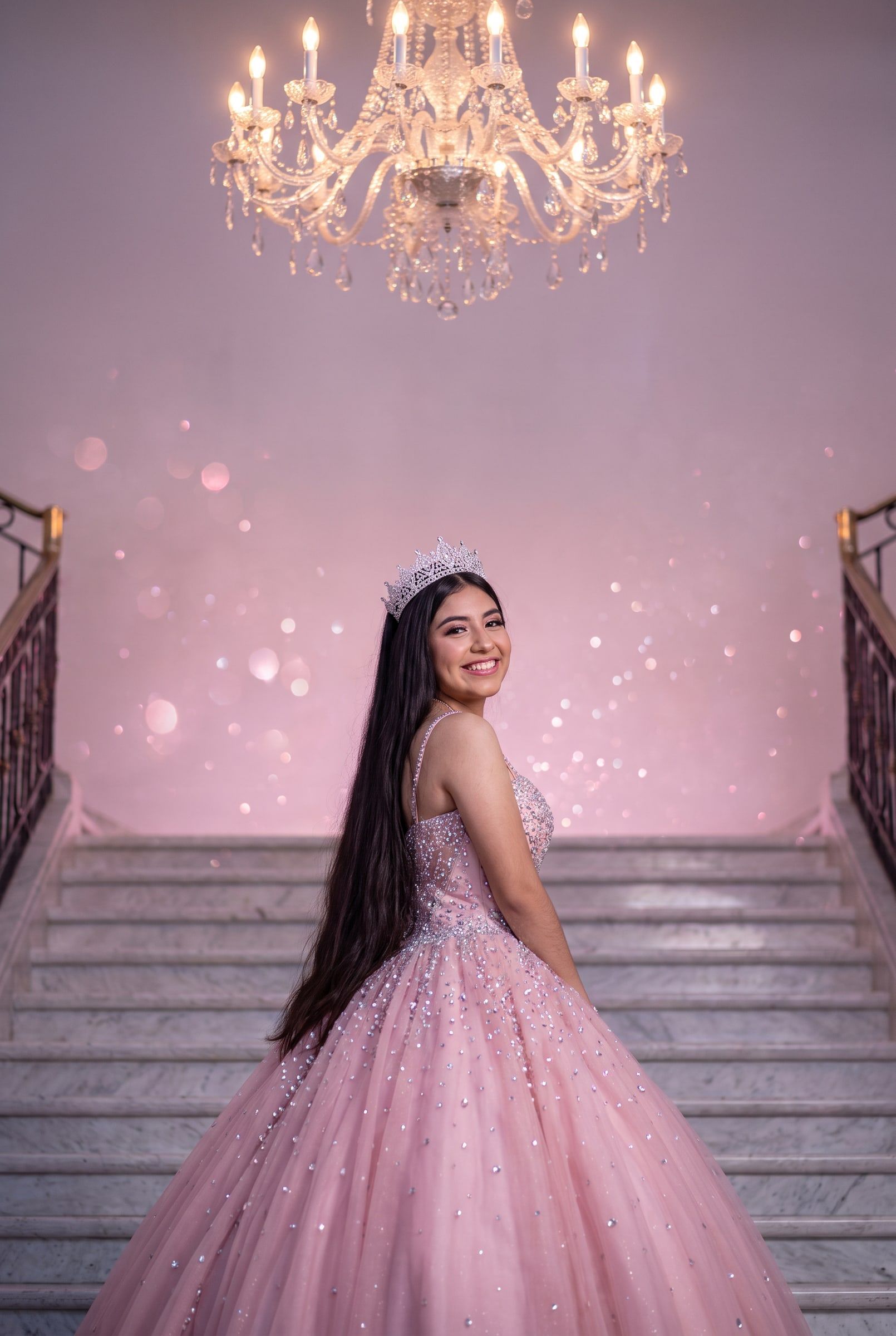 Ximena con corona plateada y vestido rosa con cristales en una escalinata de palacio